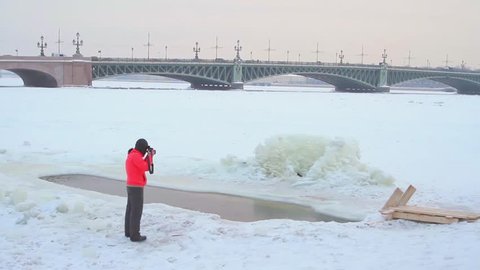  SAINT-PETERSBURG, RUSSIA -JANUARY 19, 2016: Man plunges in cold water in ice hole at winter. His photographs female photographer in a red jacket.  Trinity Bridge in the background.