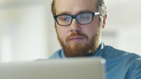 Portrait of a Bearded Young Man Wearing Glasses Sitting in His Office Working on a Computer. Computer Screen Reflects in His Glasses. Shot on RED Cinema Camera in 4K (UHD).