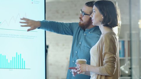 Young Man and Woman Discuss Charts Drawn on Their Electronic Whiteboard. Man Shows Details on the Screen Woman Listens Holding Cup of Coffee in Her Hands.Their Office is developer and Modern Looking.