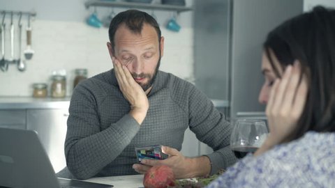 Sad, unhappy couple counting bills on smartphone and laptop in kitchen at home
