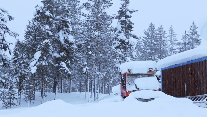On the edge of the winter forest is an old wooden house and the snow-covered tractor. Snowfall