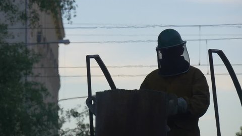 Man's Silhouette in Helmet on a Top of Furnace, Worker Puts on Protective Glasses. Male Workers Are Casting Metal Using Furnaace. Viewers in Casting Workshop Outdoors. Craftsmen From All Over the