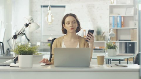 Portrait Shot of a Creative Woman Sitting at Her Desk. She Holds Smartphone and Consults Her Notebook. She Sits in a Light and Modern Office. Shot on RED Cinema Camera in 4K (UHD).