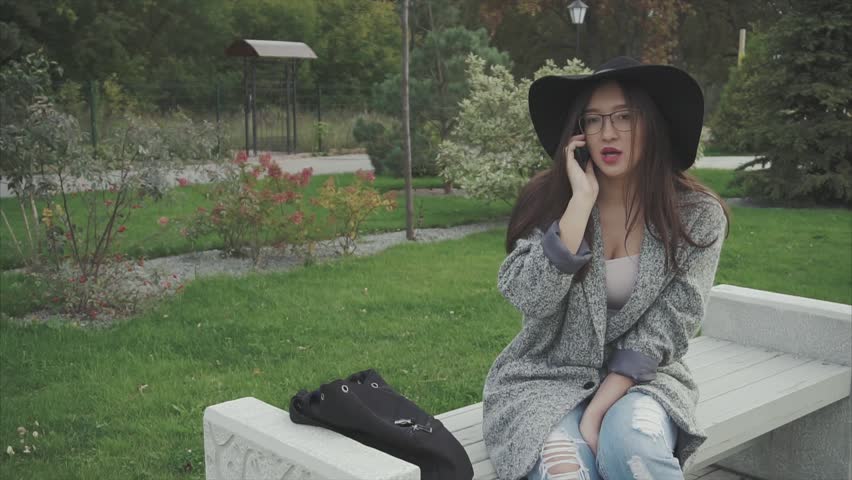 Young beautiful woman in black hat and glasses sitting on a bench in the city park and speaking on the phone and smiling