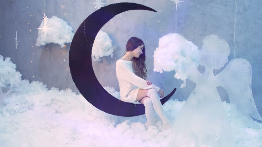 Sexy beautiful girl wearing white knited sweater over naked body singing while sitting on black moon decoration surrounded by white fluff