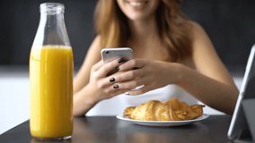 Happ young woman typing message on mobile phone while sitting on a kitchen and having breakfast
