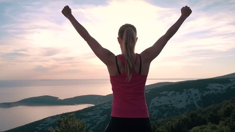 Slow motion - Young sporty female athlete raising arms victoriously after climbing the seaside mountain. Female model standing on top of the rock mountain in worship pose & celebrating successful hike