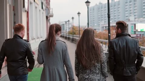 Two trendy girls and two stylish men walk in the city. Buildings at the background. Slow mo, steadicam shot, back view