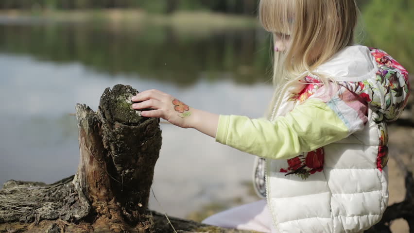 Little blonde girl bored on the lake. The child sits near the old stump. Ladybug and flower painted on children