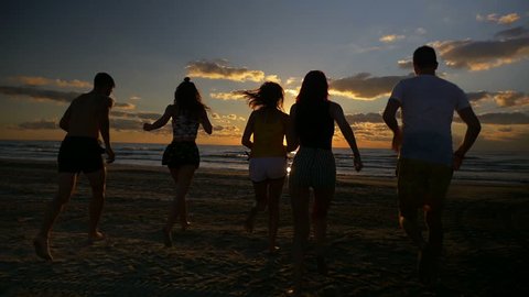Group of five friends running together towards the sea on a beach at sunset in slow motion