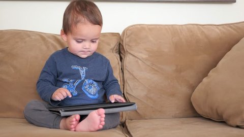 An adorable little boy plays with an iPad while sitting on a couch in his home
