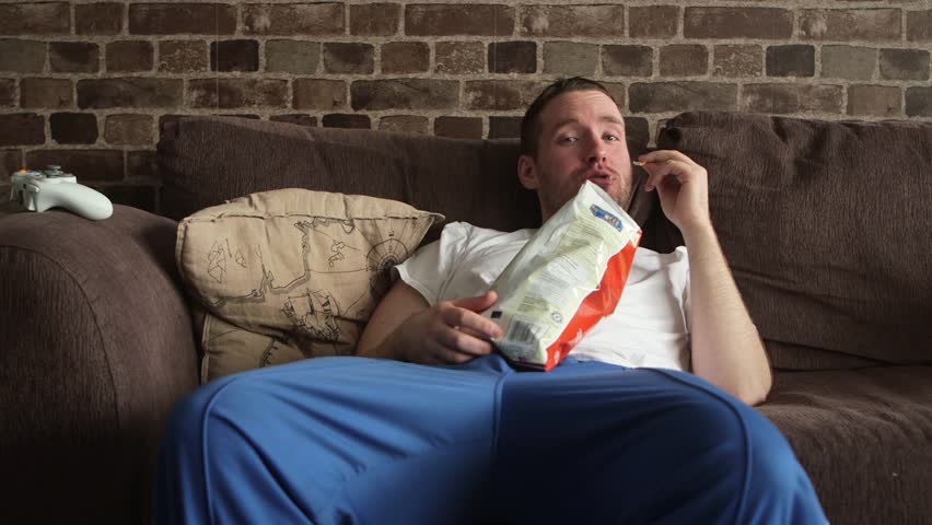 A lazy man sitting on his couch eating potato chips, watching TV and and laughing. | Shutterstock HD Video #22312957