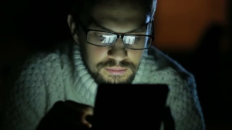 guy with glasses and a sweater with a tablet in the dark