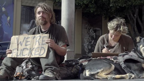 LOS ANGELES - NOV 1, 2016: Homeless Woman Writing Sign Man Holding We're Fucked Pet Dog Sitting On Street 4K Hollywood LA CA. On any given night, 82,000 people are sleeping in the street in LA County.