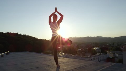SLOW MOTION CLOSE UP: Peaceful healthy young girl doing tree pose on left foot, hands in Namaste on top of skyscraper standing on edge looking down on stunning green overgrown mountains and vivid town