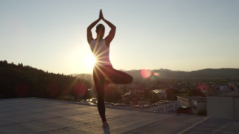 SLOW MOTION, CLOSE UP: Spiritual young girl doing tree pose on left foot, hands in Namaste on top of skyscraper standing on edge, feeling harmony and peace at golden sunset with vivid town underneath