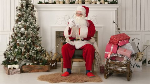 Happy Santa Claus reading Xmas messages on the phone and smiling while sitting at his chair