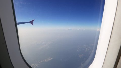 Looking out of the plane through window, small clouds blue sky, plane wing. Camera moving to the window and looking out from the plane to sky panorama above clouds at cruising altitude