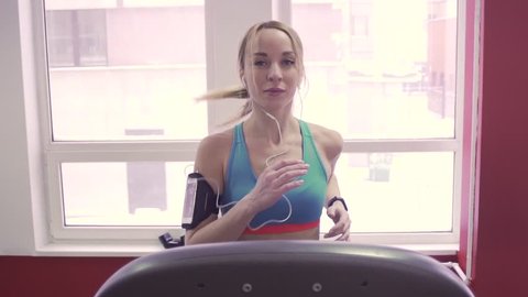 Girl running on the treadmill near a window and listening to music at the gym. Fitness lifestyle concept. Fit woman with heart rate monitor and headphones, slow motion