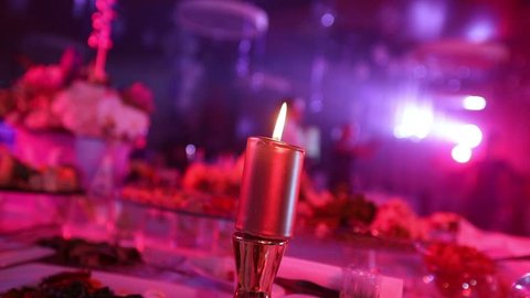 Candle on a table with a meal in the restaurant on the background of the illuminated scene, people silhouettes in the center of the restaurant hall, banquet hall, dark, shallow depth of field