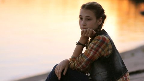 Sad girl thinking silhouette. Pretty teen girl (14-15) with braids sitting by the lake at sunrise. Pensive teenager resting chin in hand watching scenic sunset over river.