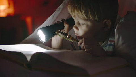 Curious little boy lying in bed in dark holding flashlight and magnifying glass and reading book