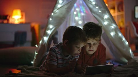 Little boys lying in teepee decorated with fairy lights and playing games on tablet in dark room