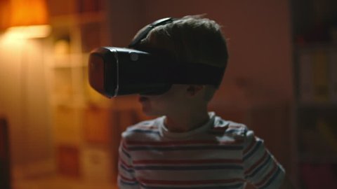 Little boy in virtual reality headset standing in dark room and looking around in amazement, ten trying to touch something invisible