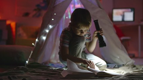 Little boy with flashlight sitting in his room near teepee decorated with fairy lights and looking at book