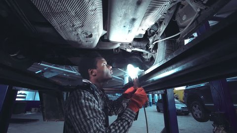 African mechanic working on the underside of a car elevated on a hoist shining a bright light onto the chassis