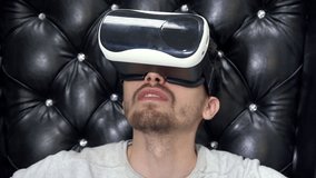 Close up shot of a young man wearing virtual reality googles on background black large leather armchair.