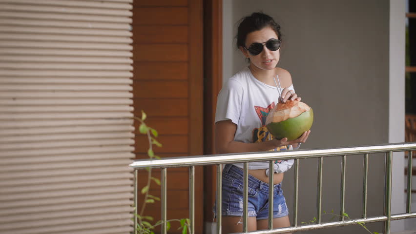 Cheerful girl with her hair pulled back in, dark glasses, dressed in a white T-shirt and short denim shorts slowly dancing on the balcony of your room and drink with relish juice from a coconut