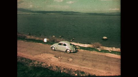 NAZARETH, ISRAEL: 1960s: VW Beetle drives along track next to coast. Motorboat on water. Sea of Galilee