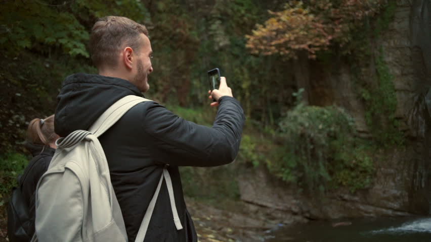 Young man taking a photo of a beautiful waterfall with a smartphone slow motion
