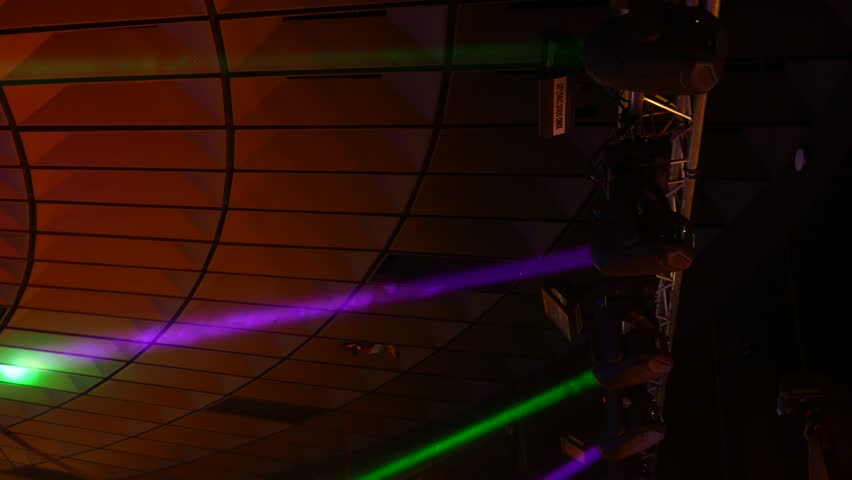 Moving light rays from row of stage illumination devices, turn and spin against concert hall roof. Dark interior, colorful beams, purple and green, automatic heads in line at ceiling rack