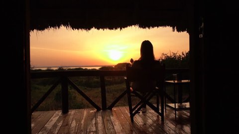 CLOSE UP: Young girl in luxury glamping Lake Burunge Tented Camp sitting on raised wooden porch, relaxing stretching legs on fence and admiring bushy landscape and beautiful golden light sunrise