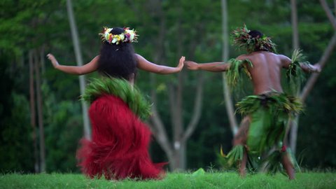 Polynesian man in warrior dress with girl in grass skirts and flower headdress dancing hula style while entertaining barefoot outdoors Tahiti French Polynesia South, Pacific,
