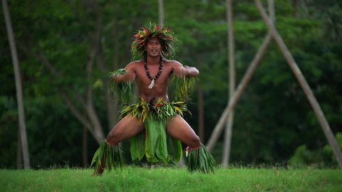 Polynesian young man in traditional grass skirts with flower headdress dancing hula war dance while entertaining barefoot outdoors Tahiti French Polynesia