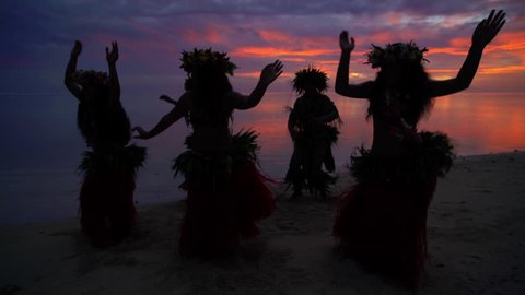 Barefoot Tahitian males in warrior dress on the beach at sunset with females in hula skirts and flower headdress performing a traditional dance French Polynesia South, Pacific,