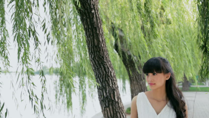 MS Young woman looking afar standing at lakeshore with willow trees in background / Beijing, China