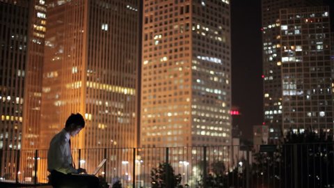 WS Businessman sitting on terrace using laptop, cityscape in background at night / Beijing, China