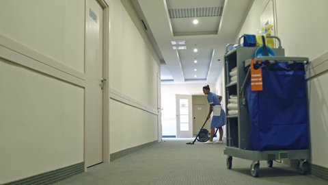 PAN of Latin American cleaning service lady in uniform vacuuming carpet in hotel hall