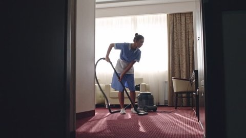 PAN of Latin American housemaid cleaning carpet in room with vacuum