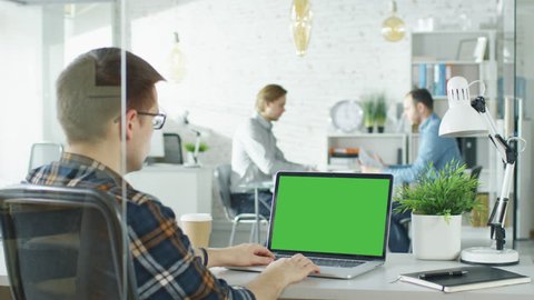 Close-up of a Man Sitting at His Desk with Green Screen Laptop. In Background Blurred and Brightly Lit Office where Two Young Man Shake Hands and Sit Down For Conversation. Shot on RED EPIC (uhd).
