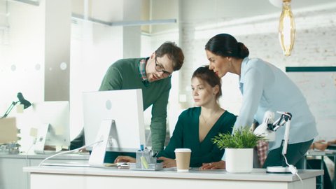 In Brightly Lit and Modern Creative Bureau. In Foreground Three People Discuss Business Issues Using Desktop Computer. In Background Groups of Coworkers Discuss Work. Shot on RED EPIC (uhd).