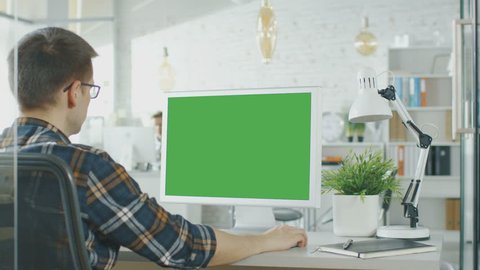 Close-up of a Man Sitting at His Desk with Green Screen PC on the Table. In Background Blurred and Brightly Lit Office where People go Through Office Routine. Shot on RED EPIC (uhd).