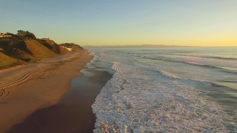 Aerial flying shot down a beach in California over the sand and water. Beach houses in the distance. Sunset time with clear blue skies and golden light.