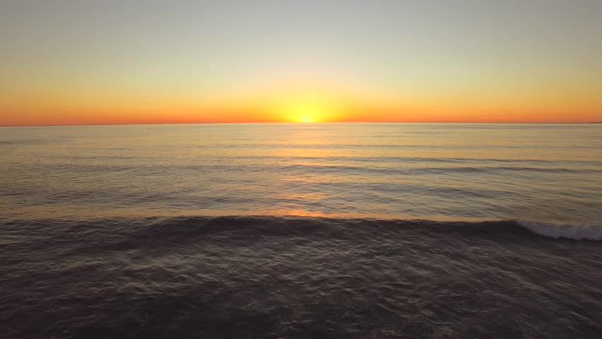 Aerial shot flying backwards over the ocean in California during sunset. Sharp horizon line with golden light. Rises at the to end reveal beach and waves. Clear skies, vibrant colors.