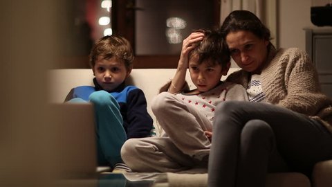 Mother and children watching TV screen at the living room. Mom with concerned look in her face worried about the content they are watching