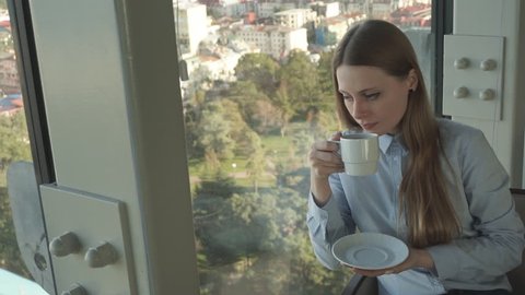 Concept: millennial, connections, lifestyle. Business casual dressed attractive girl drink tea by the window of trendy restaurant in skyscraper. HD 1080p.